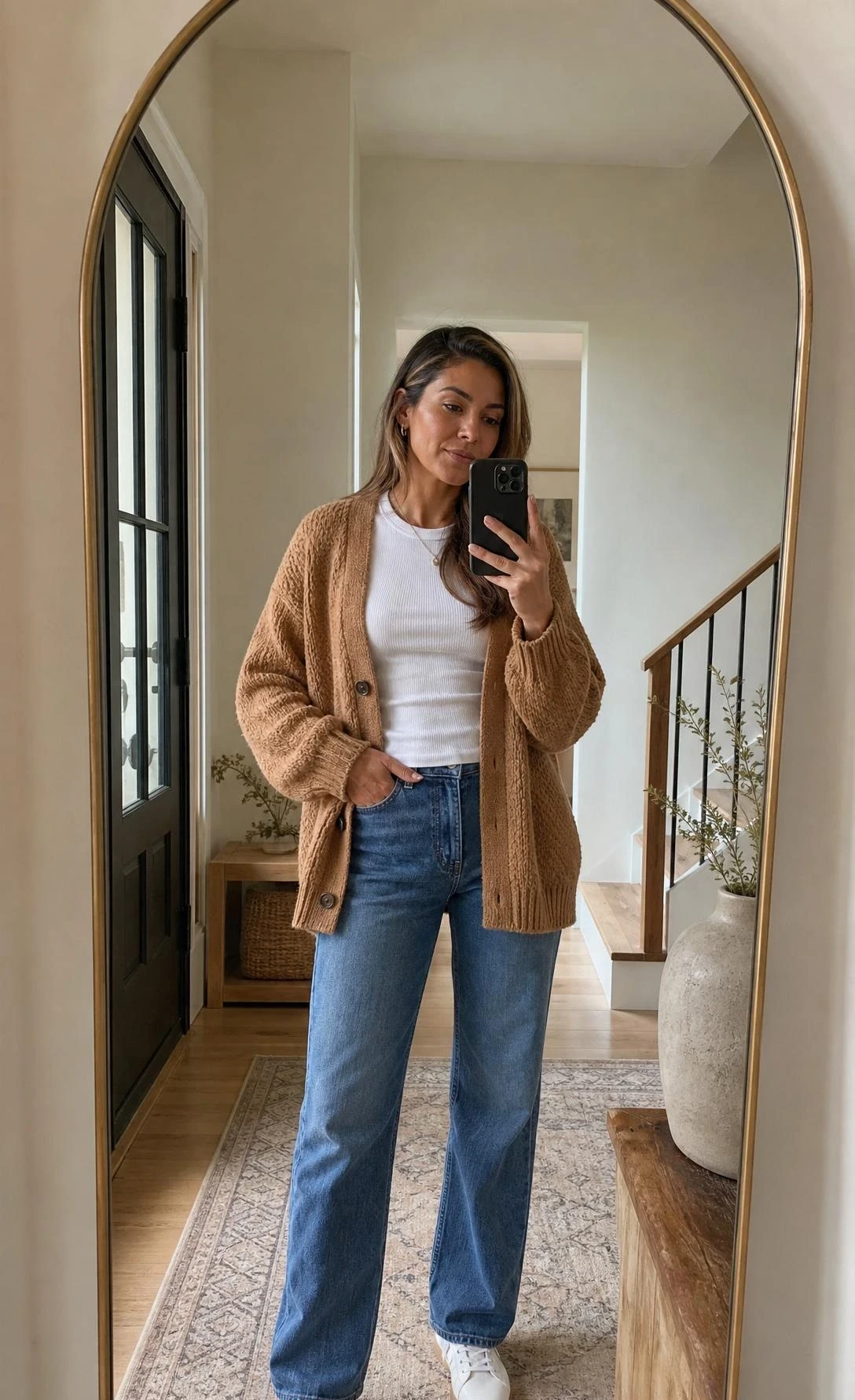 Woman in chunky-knit camel cardigan over white ribbed tee and straight jeans takes mirror selfie in warm home entryway — effortless neutral outfit