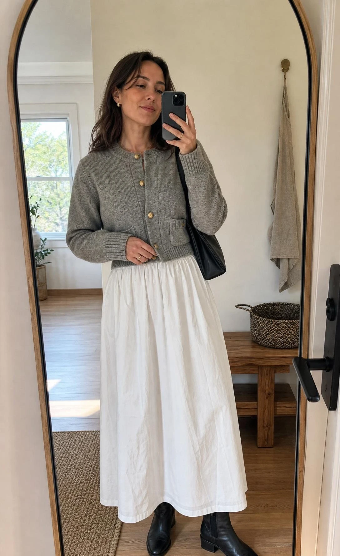 Mirror selfie of a woman styling white maxi skirt outfit ideas with a cardigan jacket and ankle boots in a clean entryway