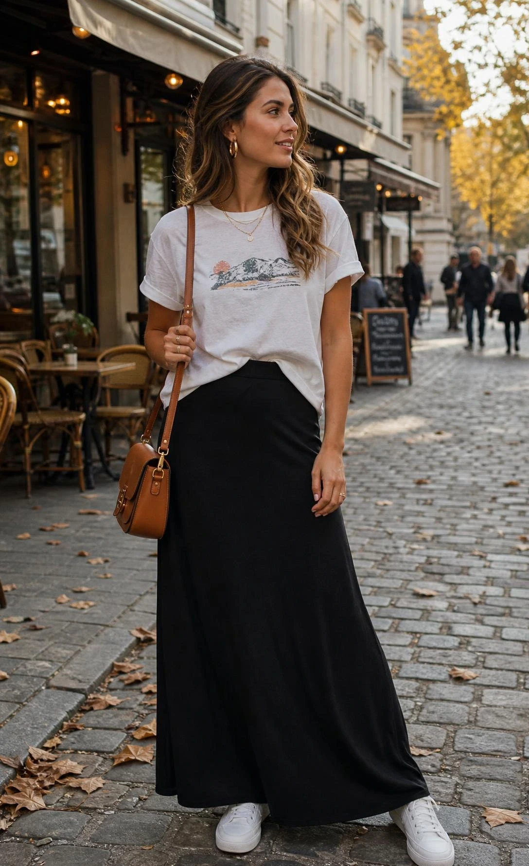 Woman half-tucking a faded graphic tee into a flowing black maxi skirt on a sun-lit cobblestone street, effortless casual style