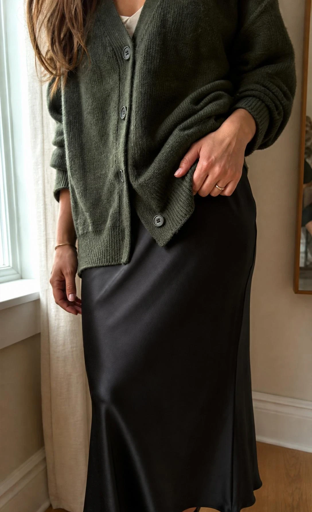 Close-up of a woman by a bright window wearing a forest green cardigan with a black slip skirt, highlighting rich knit texture and sleek contrast.