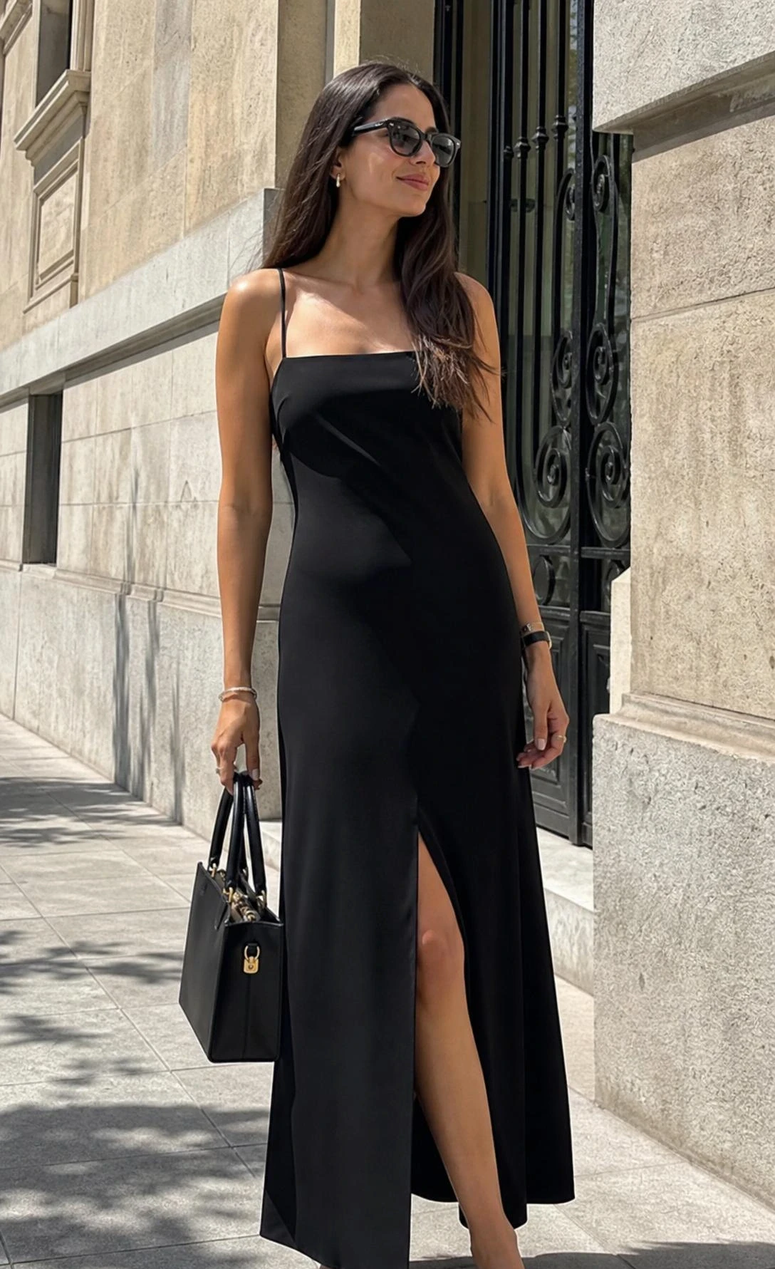 Low-angle street style shot of a tall slim woman in a black maxi dress with slit and structured accessories.