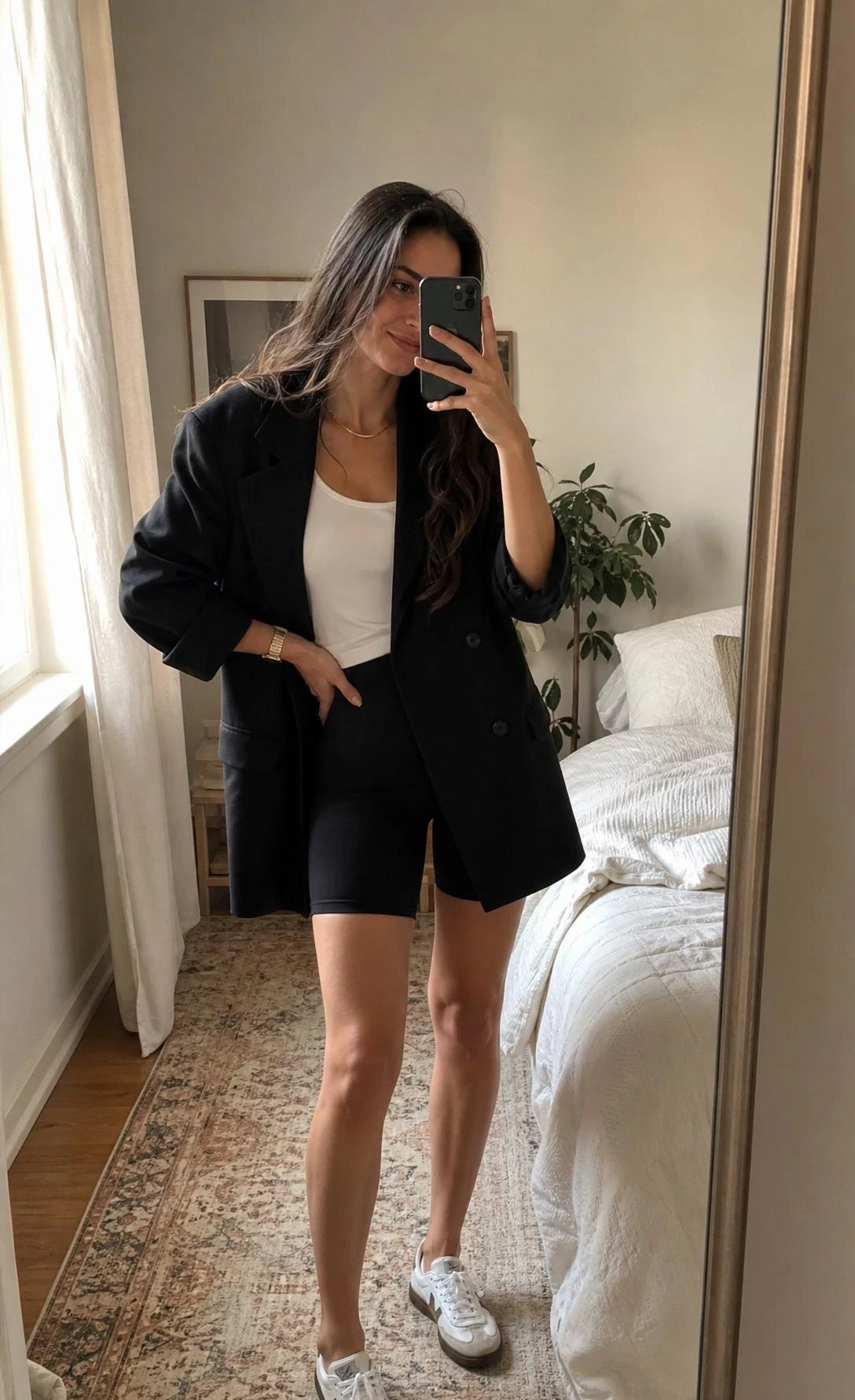 Mirror selfie of a tall slim woman in an oversized blazer, biker shorts, and white tank in a softly lit bedroom.