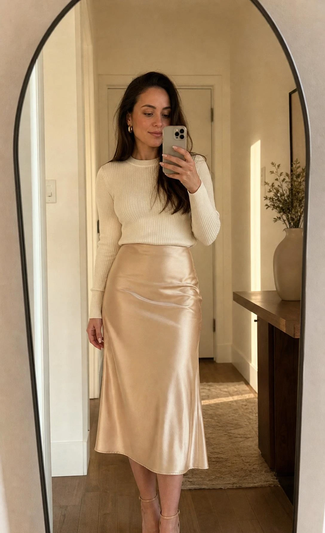 Woman styling a satin midi skirt with a fitted ribbed knit top for a cocktail outfit, captured in a mirror selfie in a softly lit home entryway