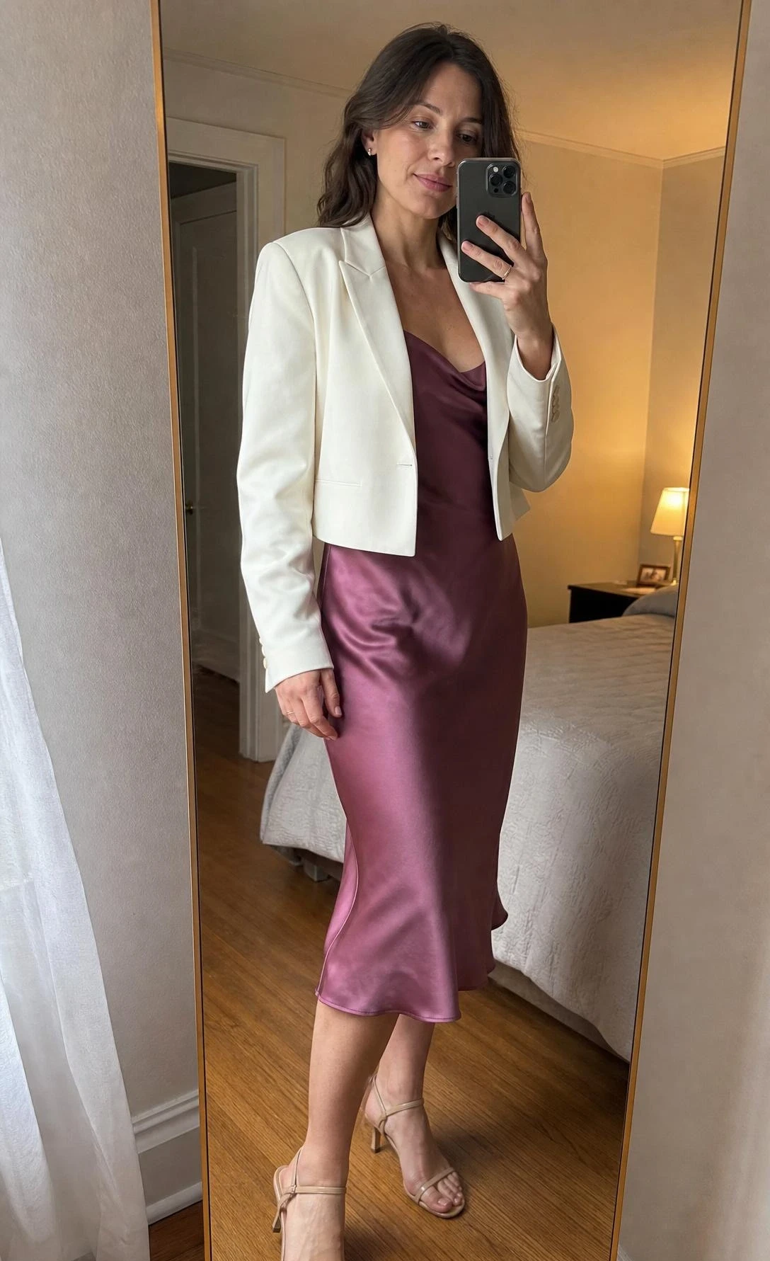 Woman in slip dress with cropped structured jacket poses for mirror selfie in warmly lit bedroom, showing a polished cocktail outfit combination