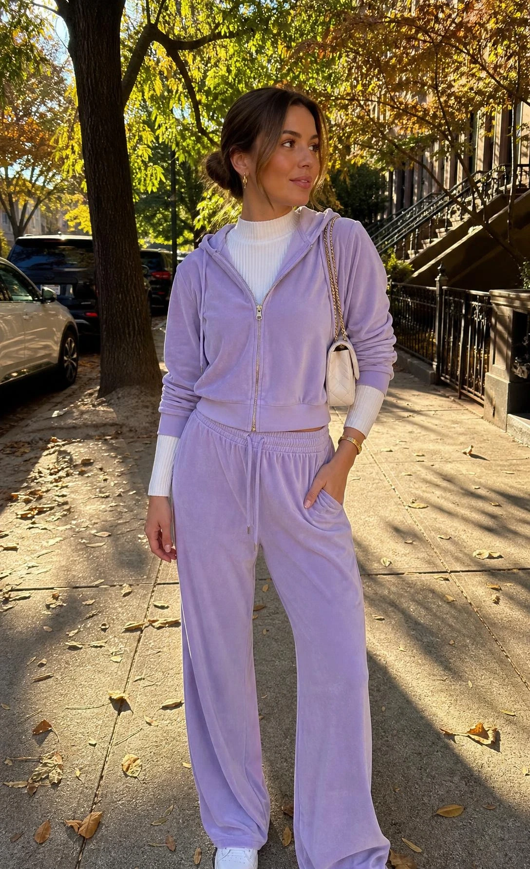 Woman wearing a lavender velour tracksuit on a tree-lined sidewalk, capturing the iconic Y2K outfit trend in a soft pastel hue