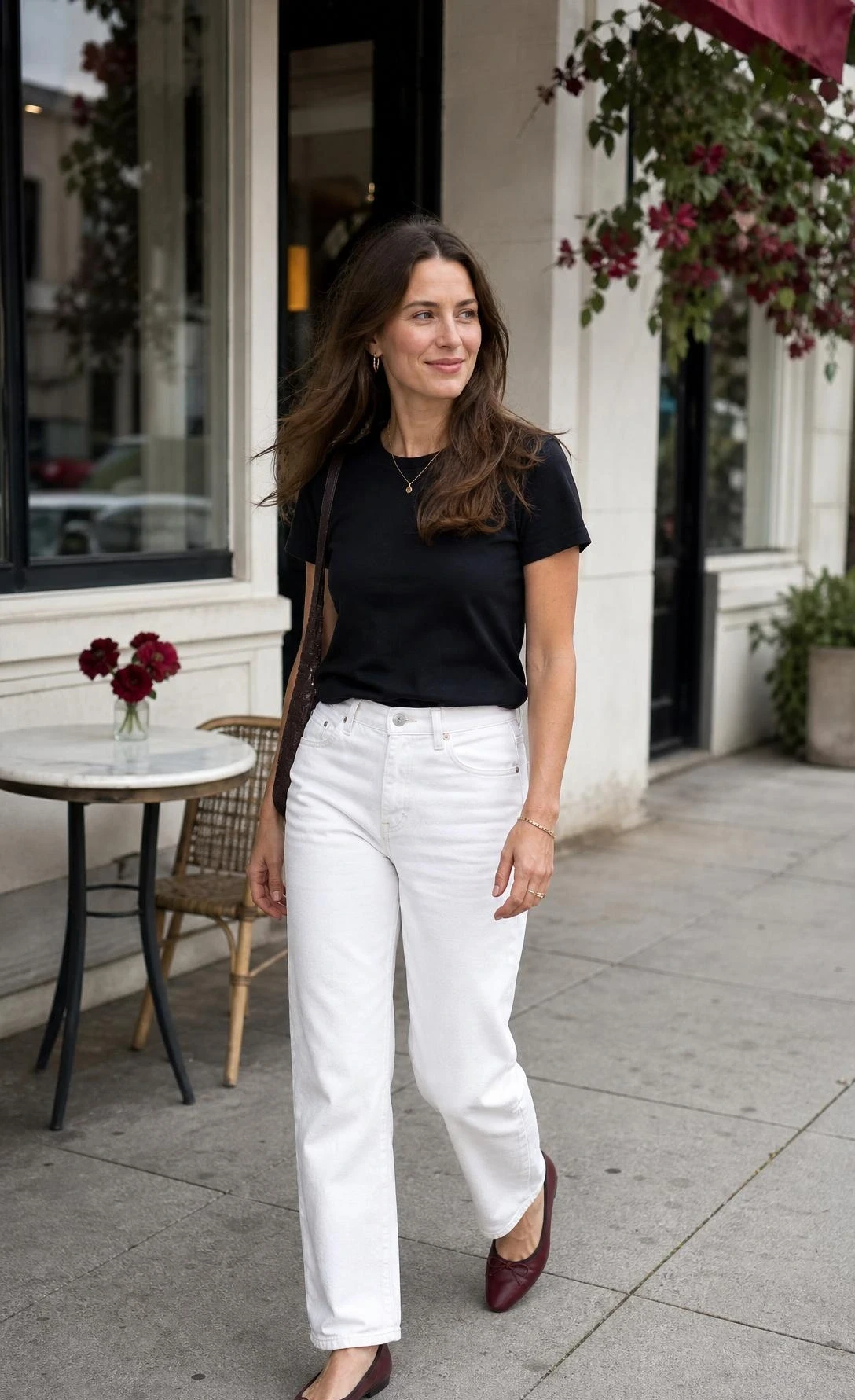 Woman walking outside a cafe in one of the white jeans outfit ideas: a black tucked tee and burgundy flats