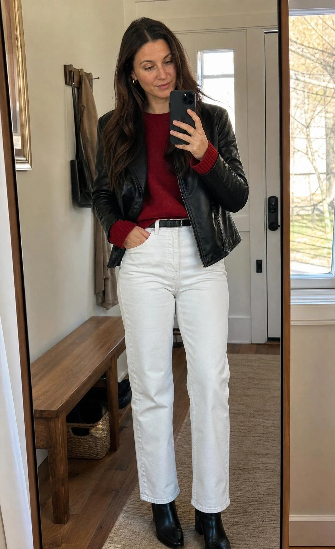 Woman takes an entryway mirror selfie in a red sweater, black leather jacket, and white jeans outfit ideas look