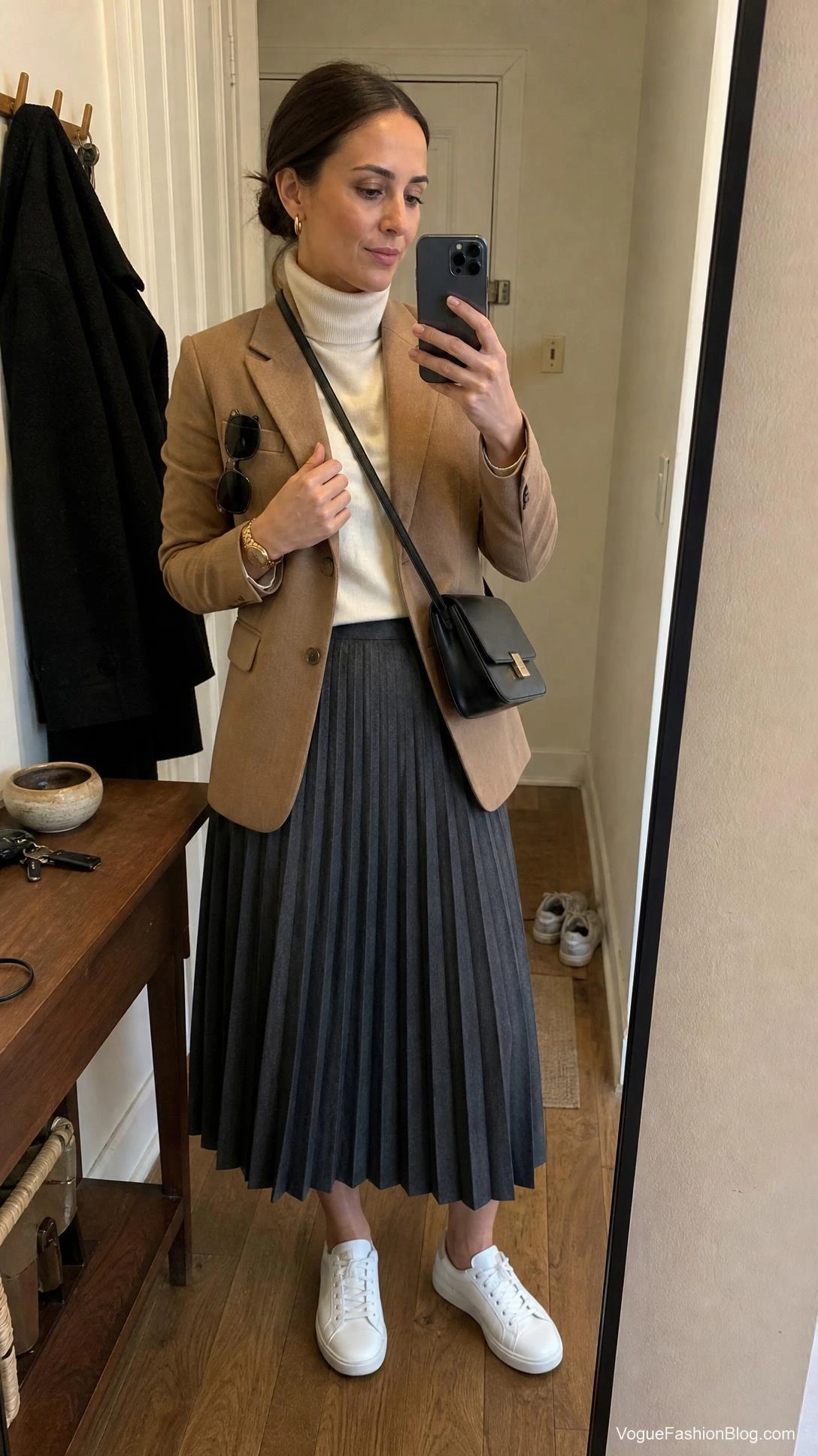 Woman in camel blazer and cream turtleneck styling a pleated midi skirt outfit for fall with white sneakers in an entry mirror.