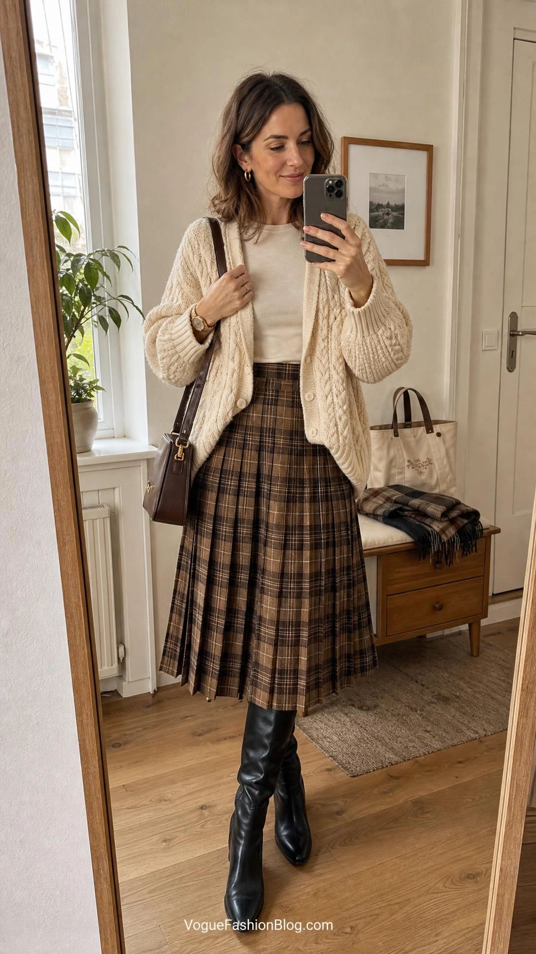 Mirror selfie of a fall pleated midi skirt outfit with chunky cream cardigan, ivory top, and muted plaid skirt