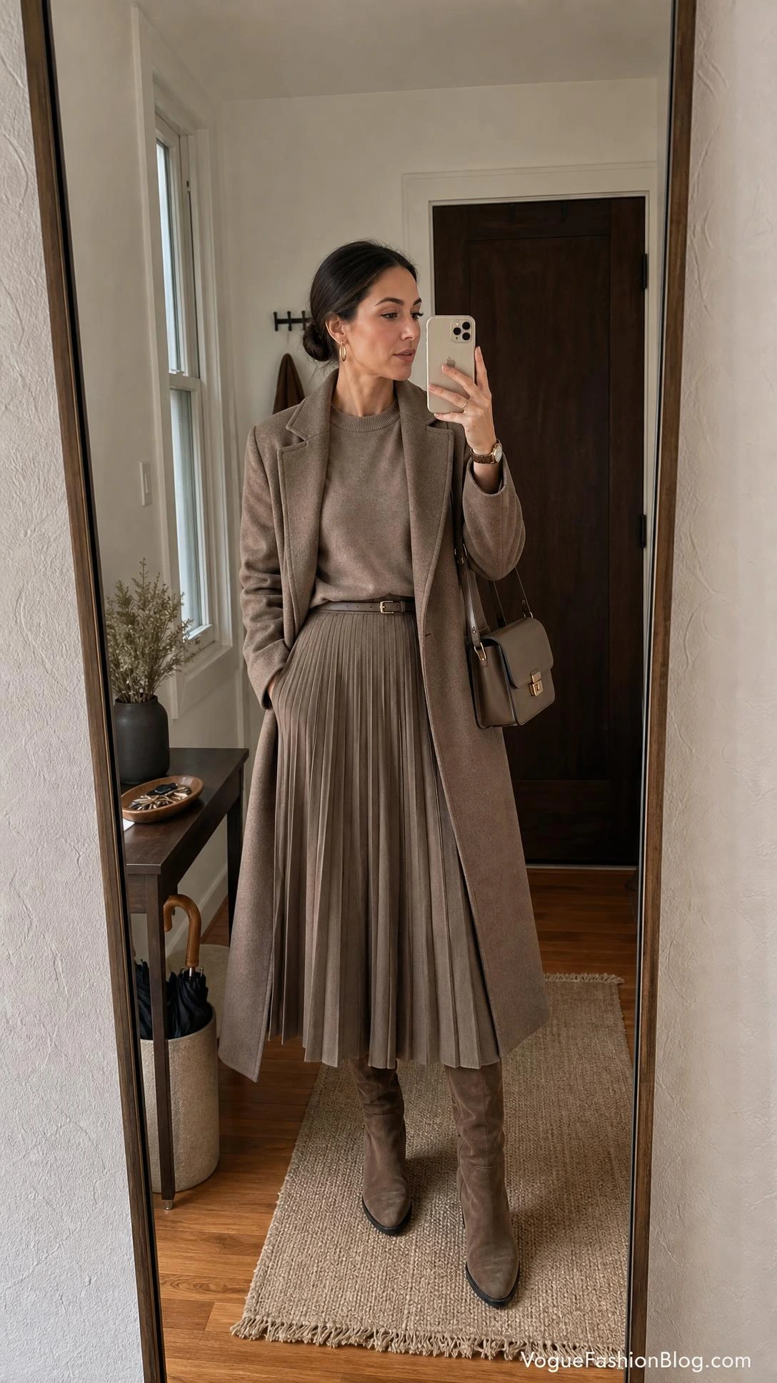 Woman takes entryway mirror selfie in a long wool coat over a monochrome pleated midi skirt outfit for fall styling.