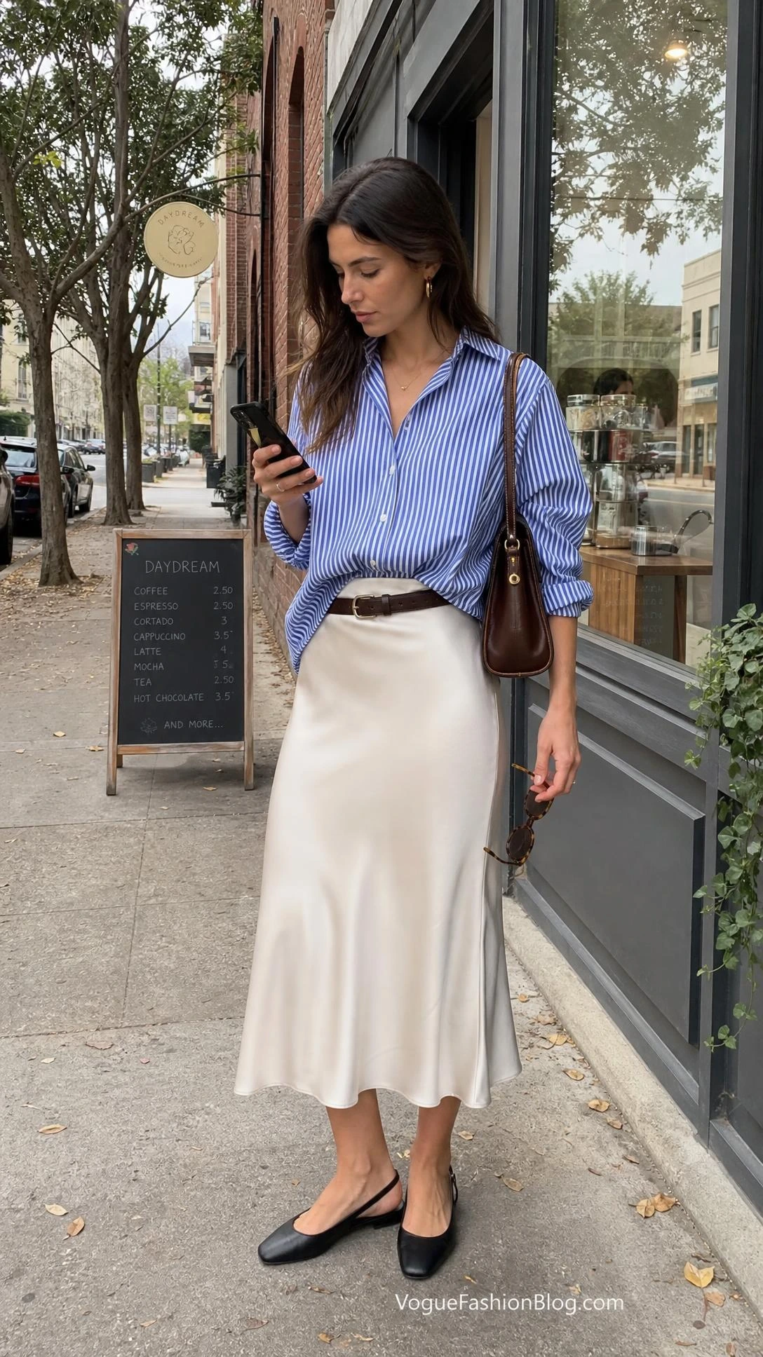 Woman outside a café shows how striped shirt outfits work with an oversized blue-and-white button-down and ivory satin slip skirt