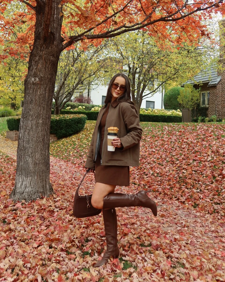 Brown matching jacket and skirt set with tall boots styled among vibrant autumn leaves.