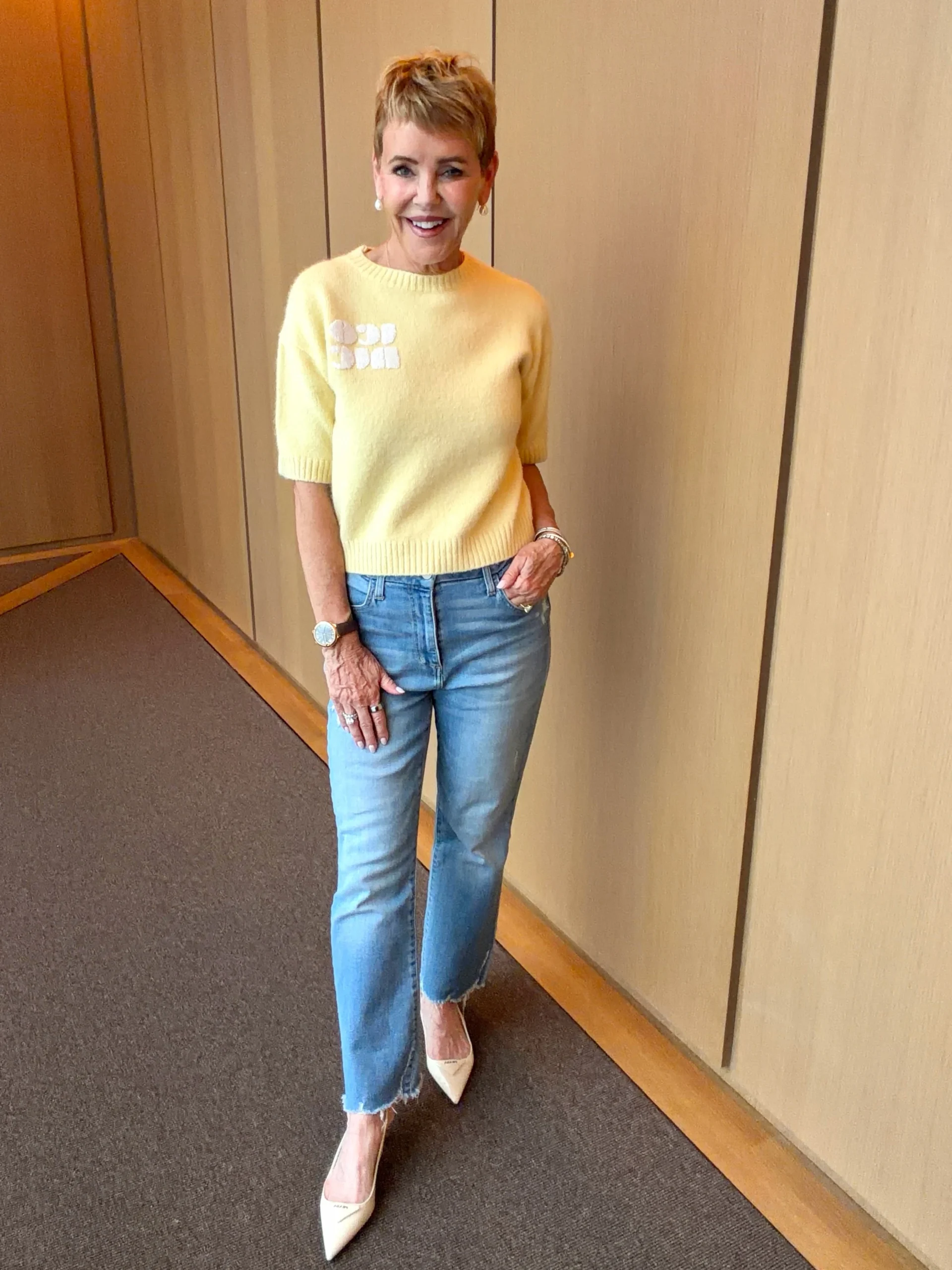 Woman wearing a mustard yellow sweater, light blue straight-leg jeans, tan belt, pearl earrings, and bracelets.