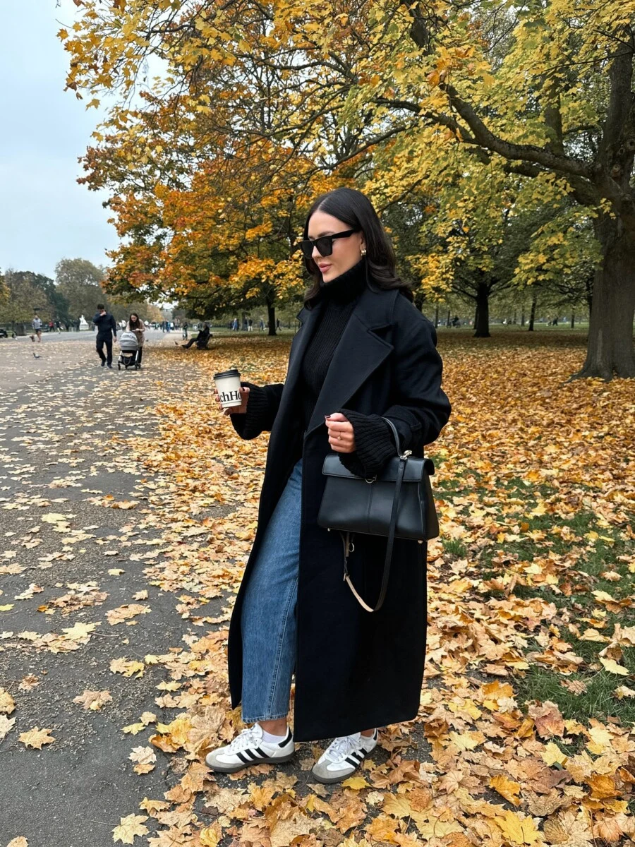 Long black coat worn over jeans and sneakers with coffee in hand for a minimal fall look.