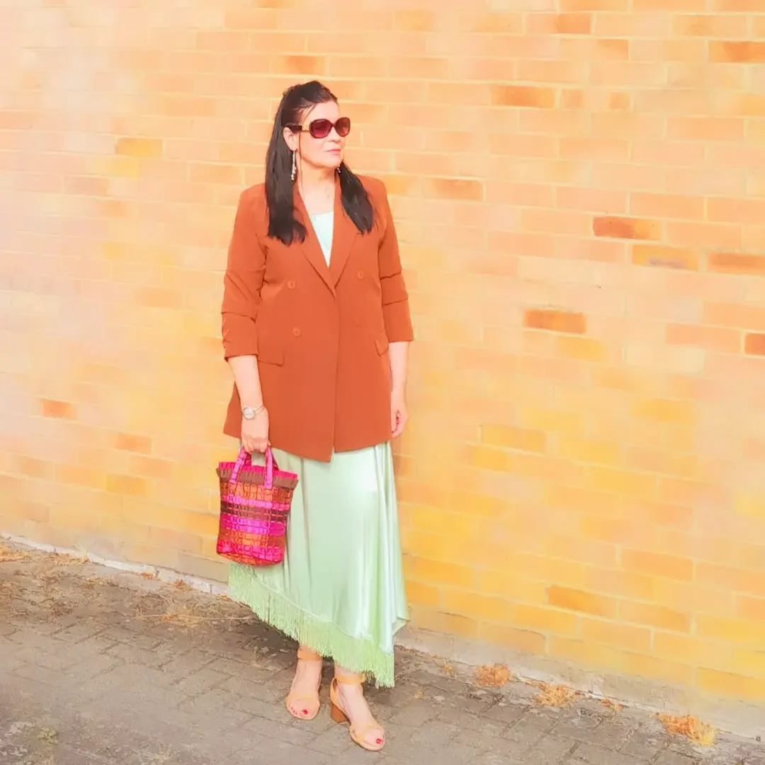 Woman in a rust orange blazer, mint green pleated midi skirt, white top, dark sunglasses, and gold watch.