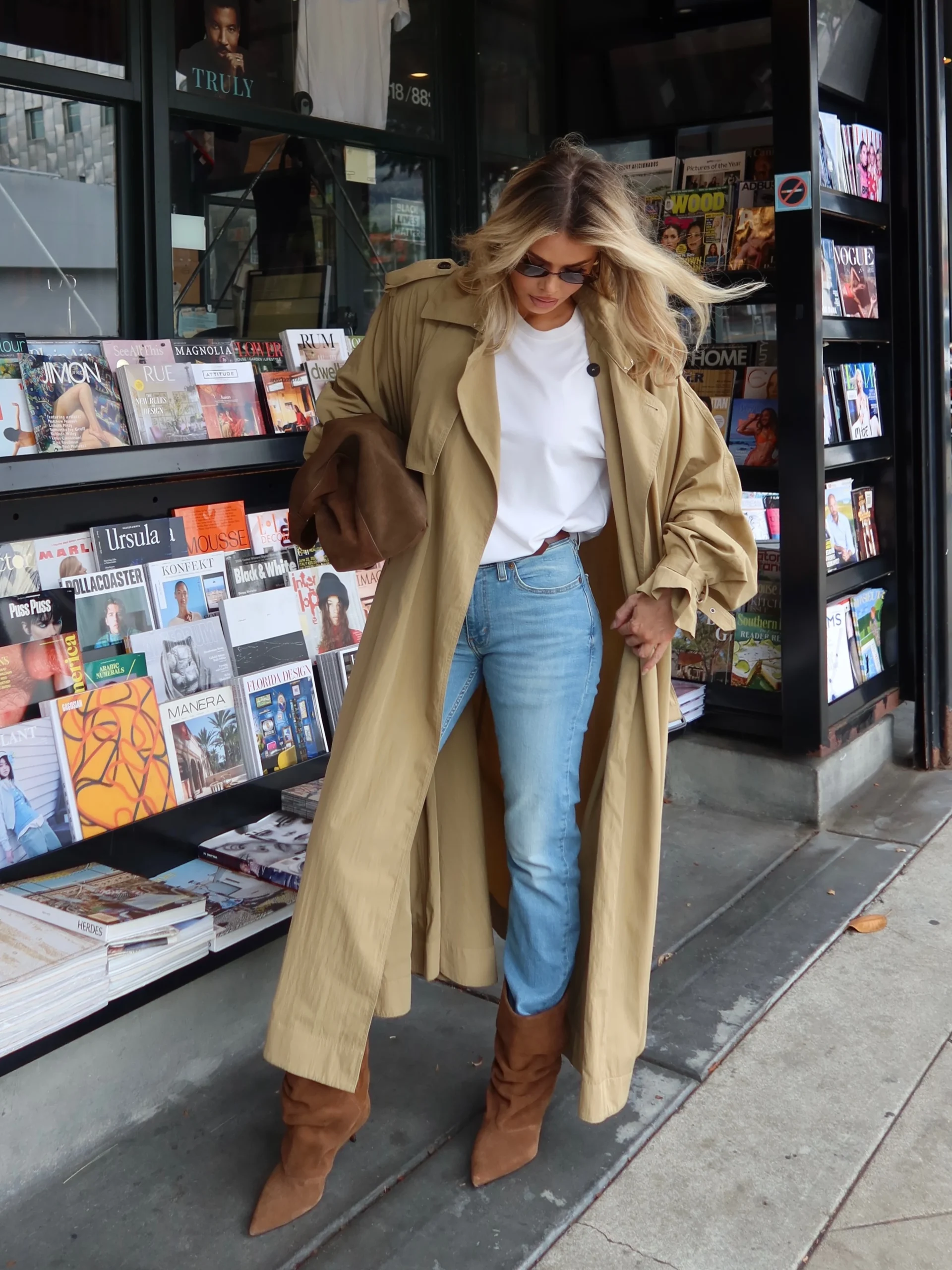 Woman in beige trench, white tee, light wash Levi's 501s, brown suede boots and oval sunglasses.