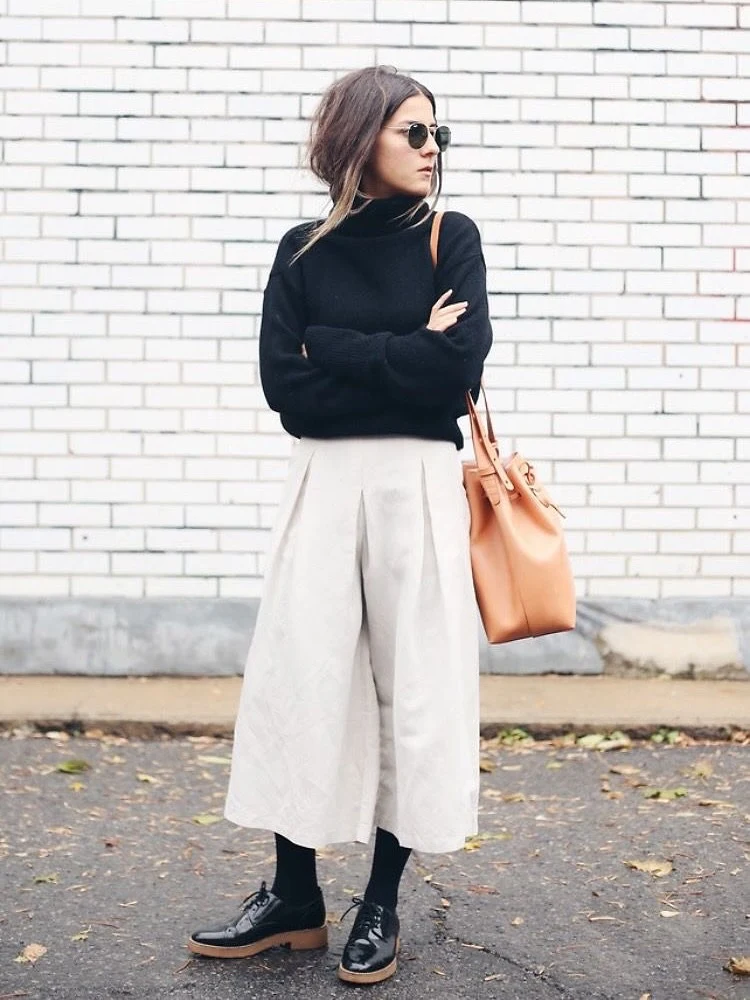 Black turtleneck and cream culottes with oxford shoes posed against a brick wall.