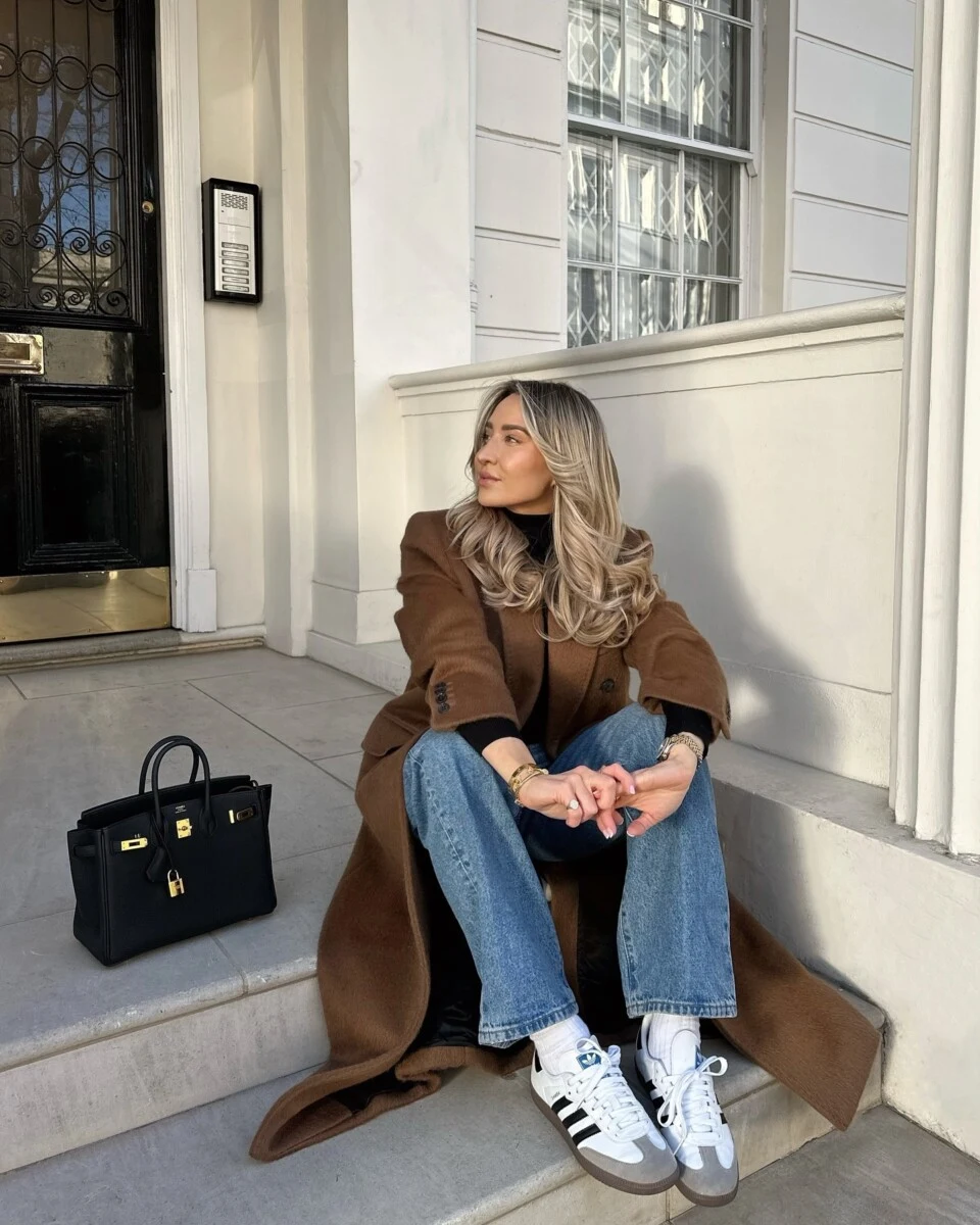 Long brown coat styled over jeans and white sneakers on a casual fall doorstep.
