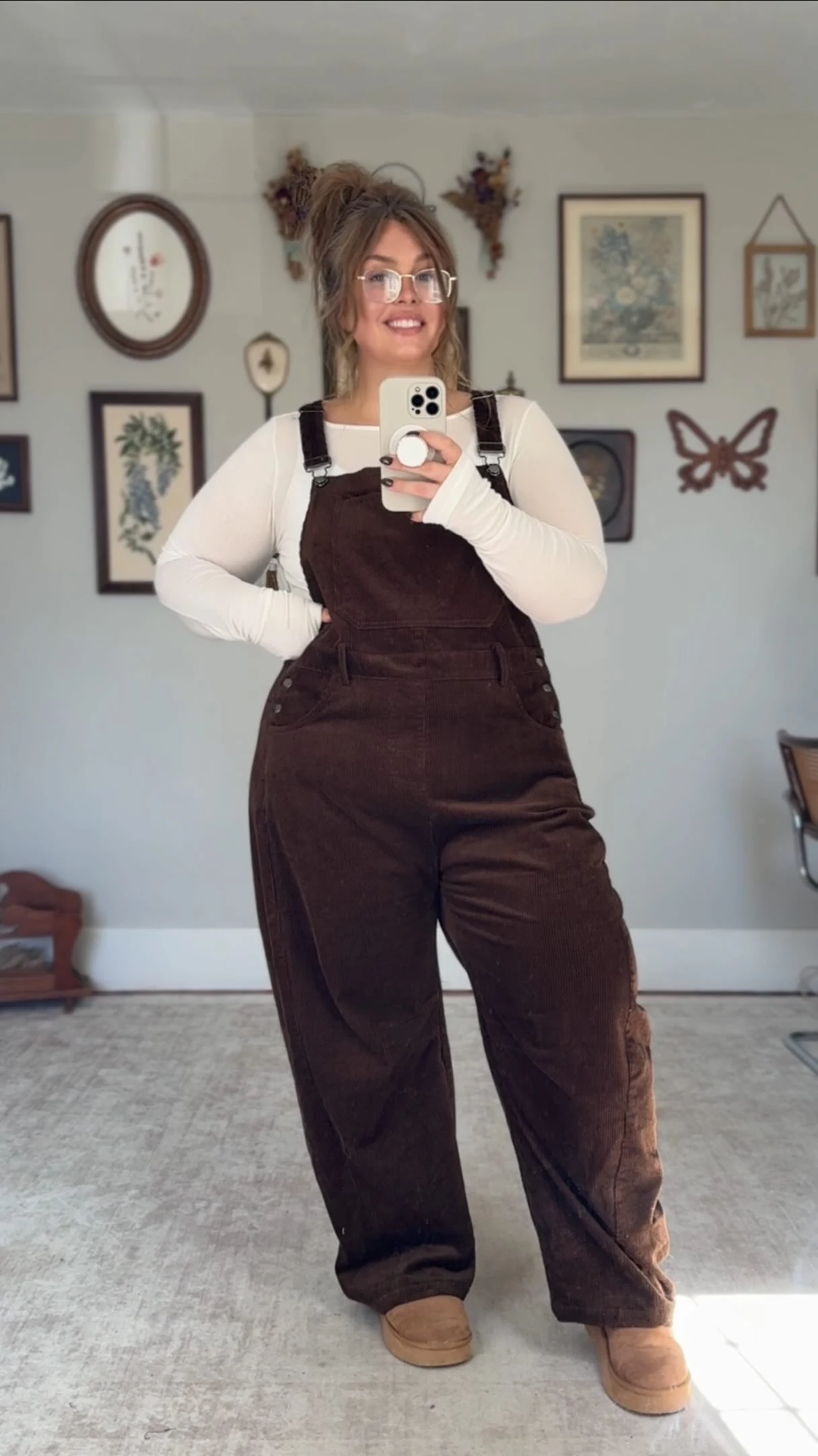 Plus size woman in brown corduroy overalls layered over a white long sleeve shirt with tan slip-on shoes.