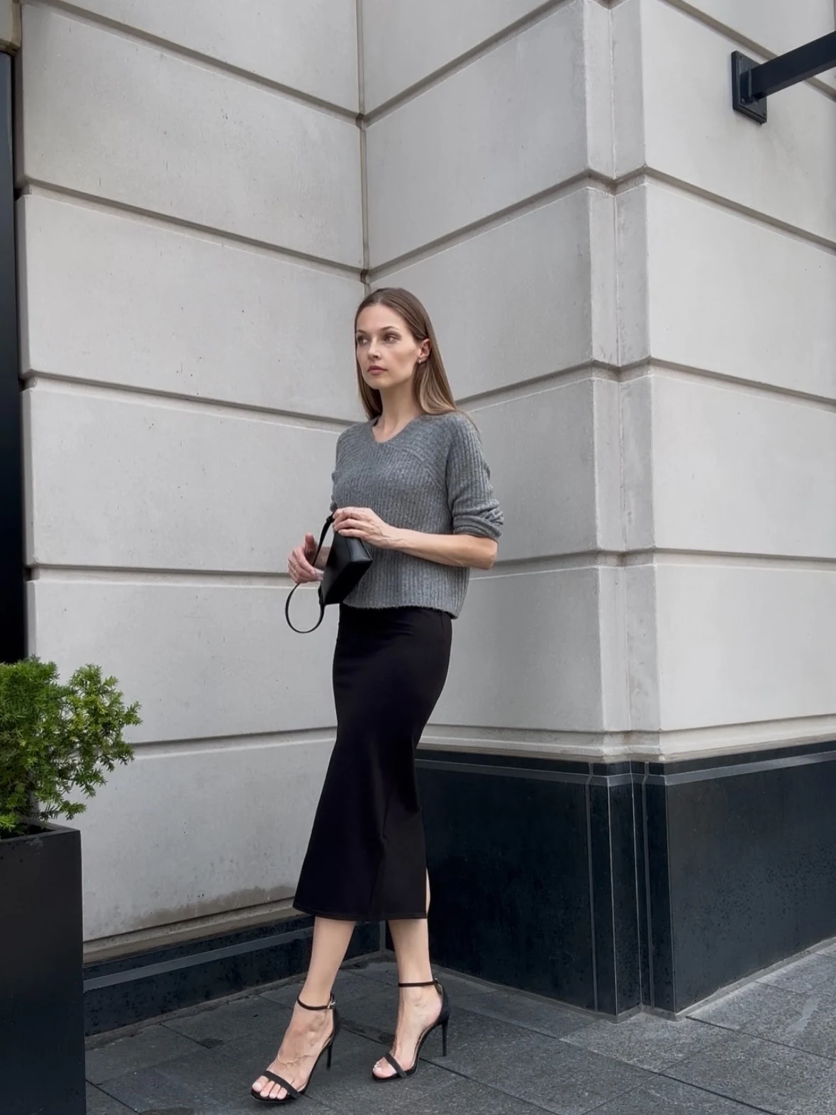 Woman in gray ribbed sweater, black satin midi skirt, and strappy heels standing outside a building.