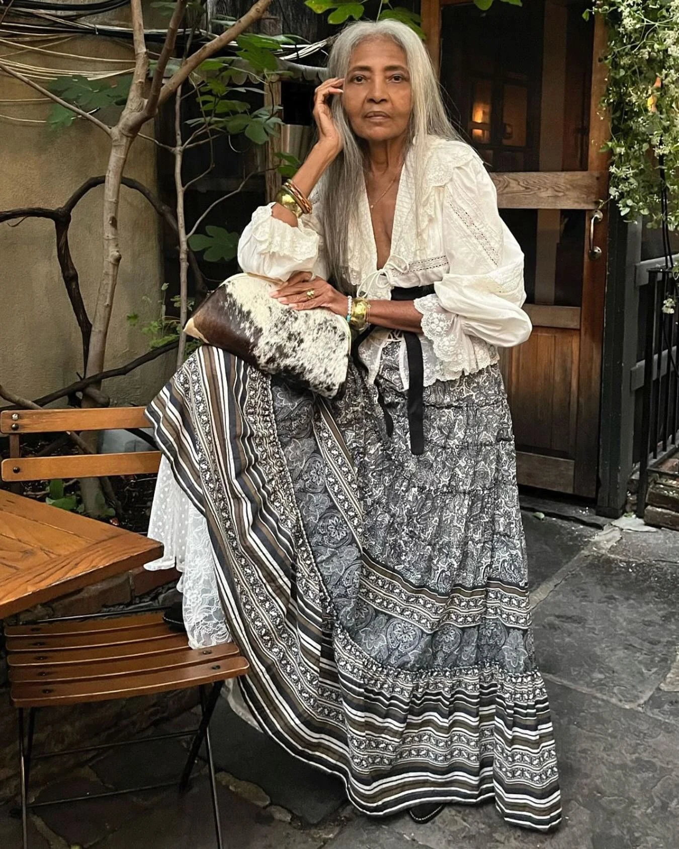 white lace-trimmed blouse with tie front, long black and white patterned maxi skirt, gold bracelets, cowhide clutch