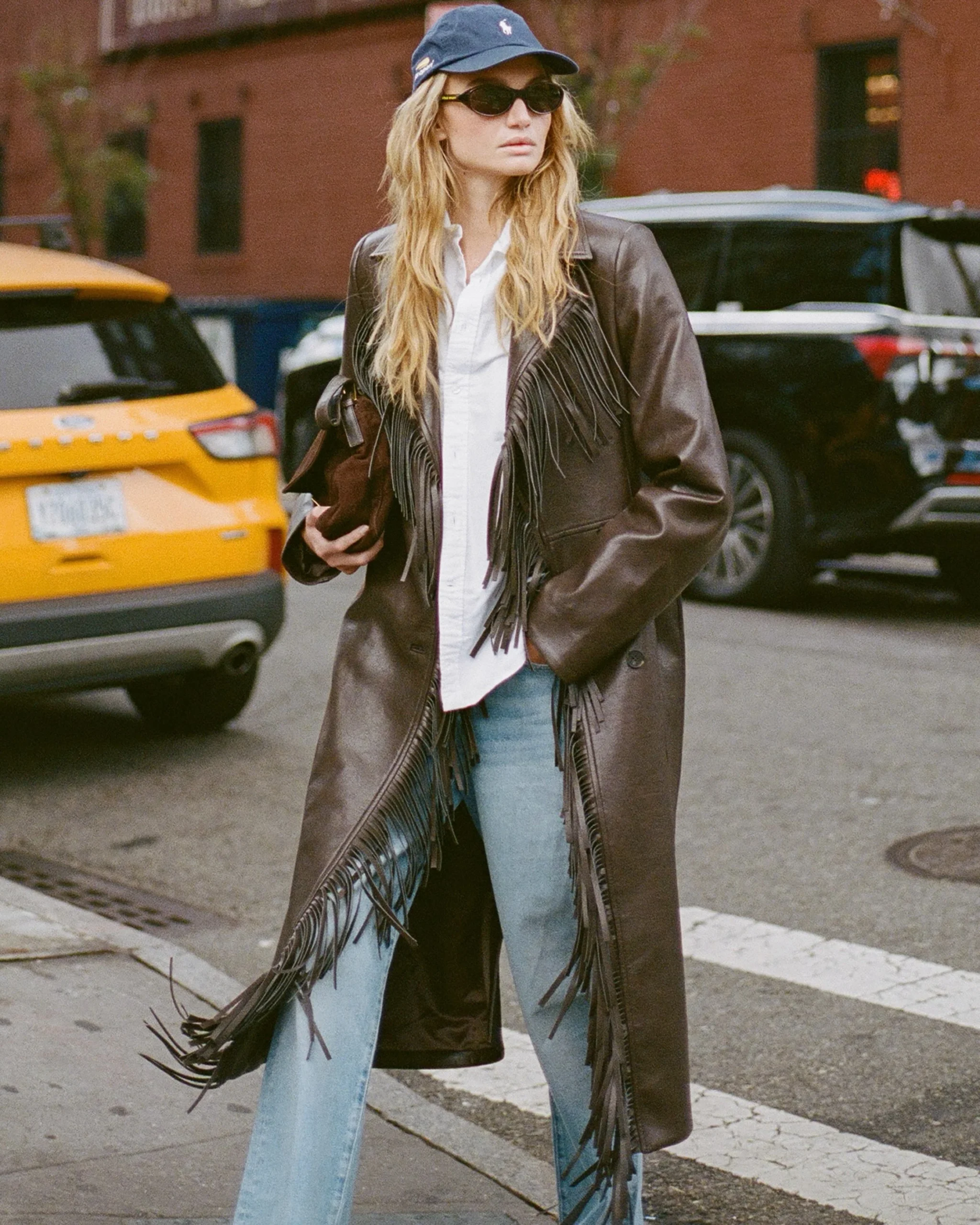 Fringe brown leather coat over white shirt and jeans with a baseball cap and suede clutch.