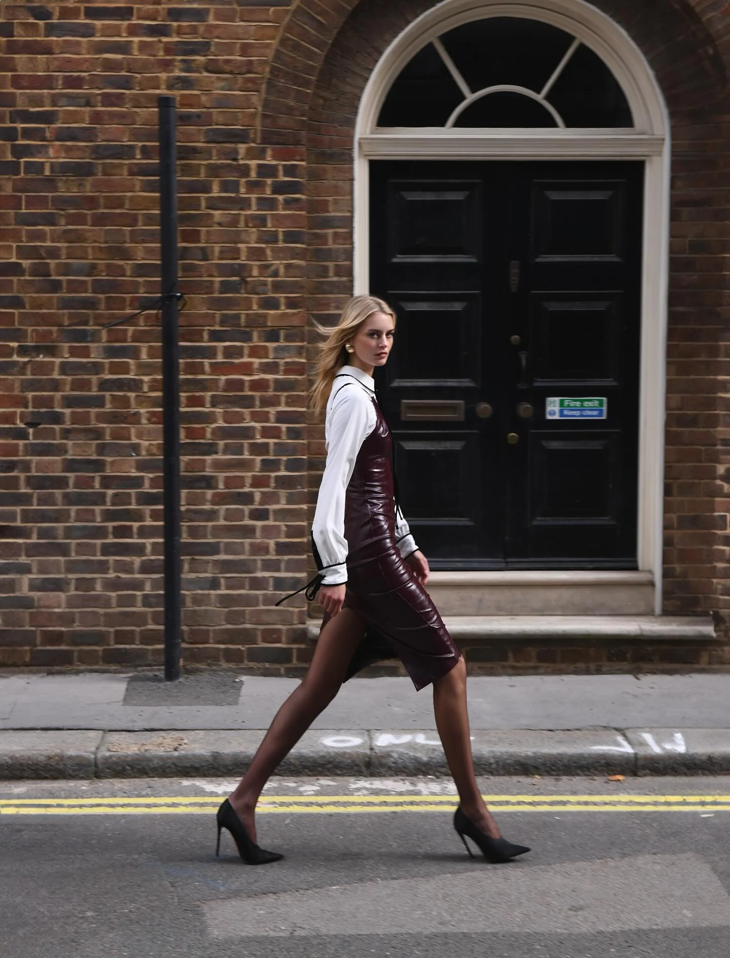 Burgundy leather-look midi dress layered over a white collared shirt with sheer tights and black stilettos.
