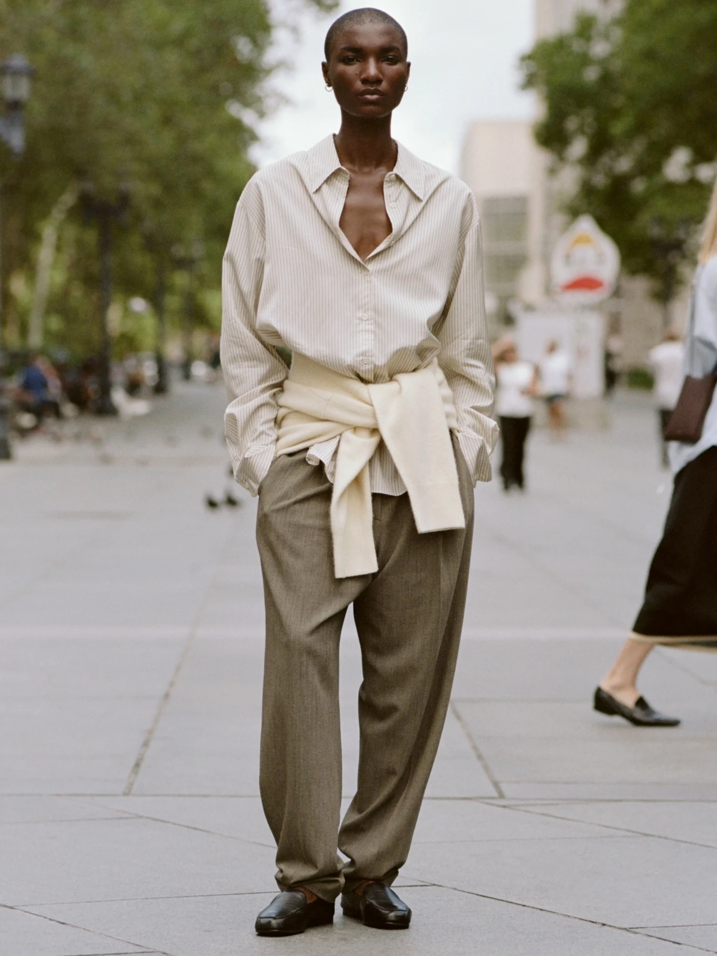 Oversized pinstripe shirt open at top, loose gray trousers, black loafers, cream sweater tied at waist.