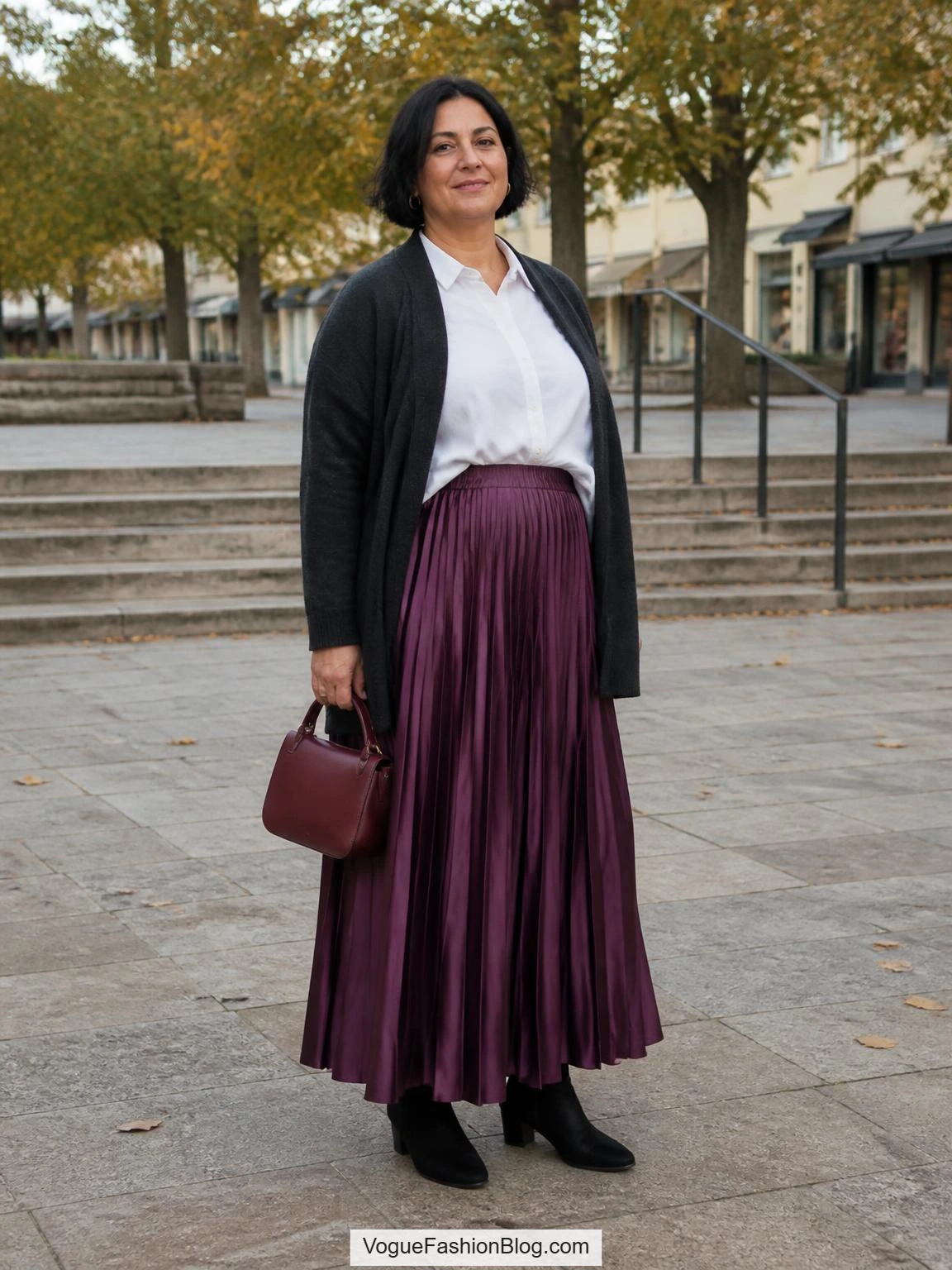 A plus-size woman styles a plum pleated satin skirt, white shirt, charcoal cardigan, and ankle boots in a fall town square.