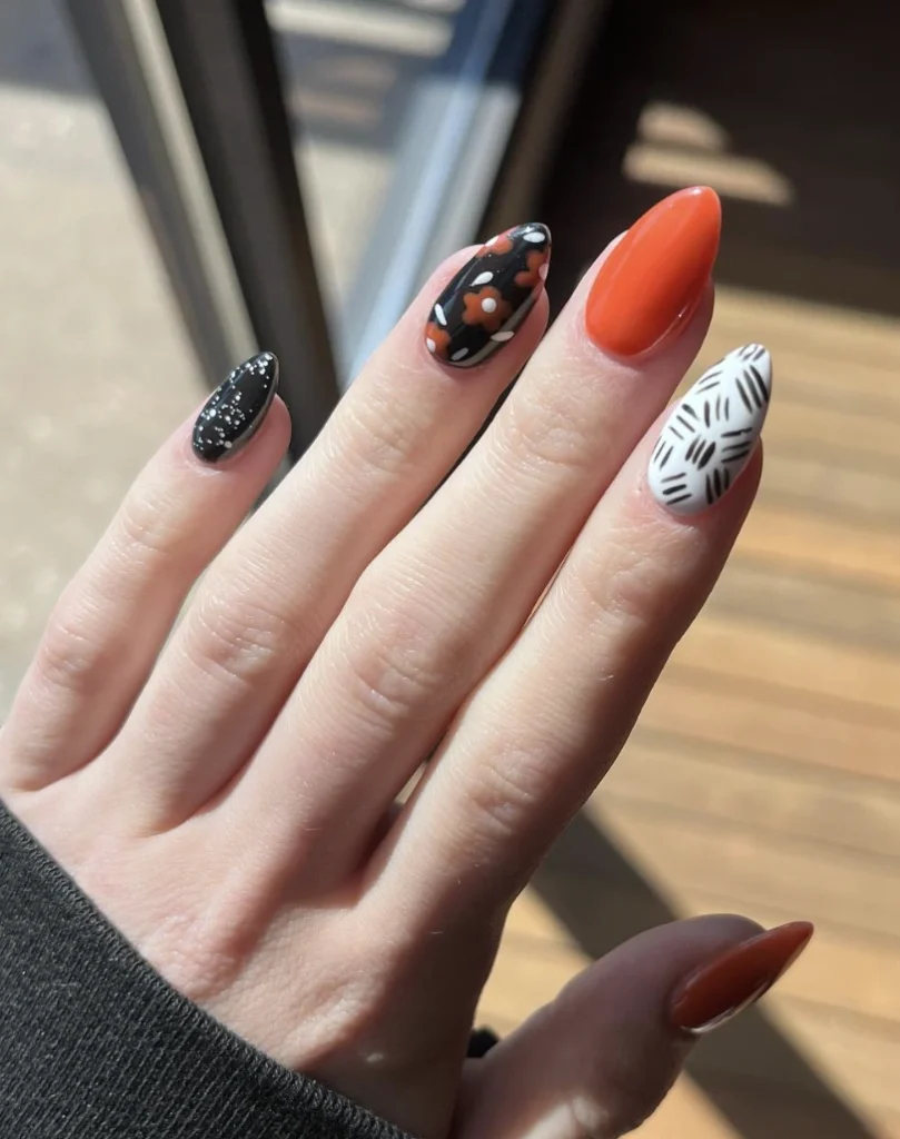 Stiletto nails in glossy black, orange, and white, featuring floral and geometric designs